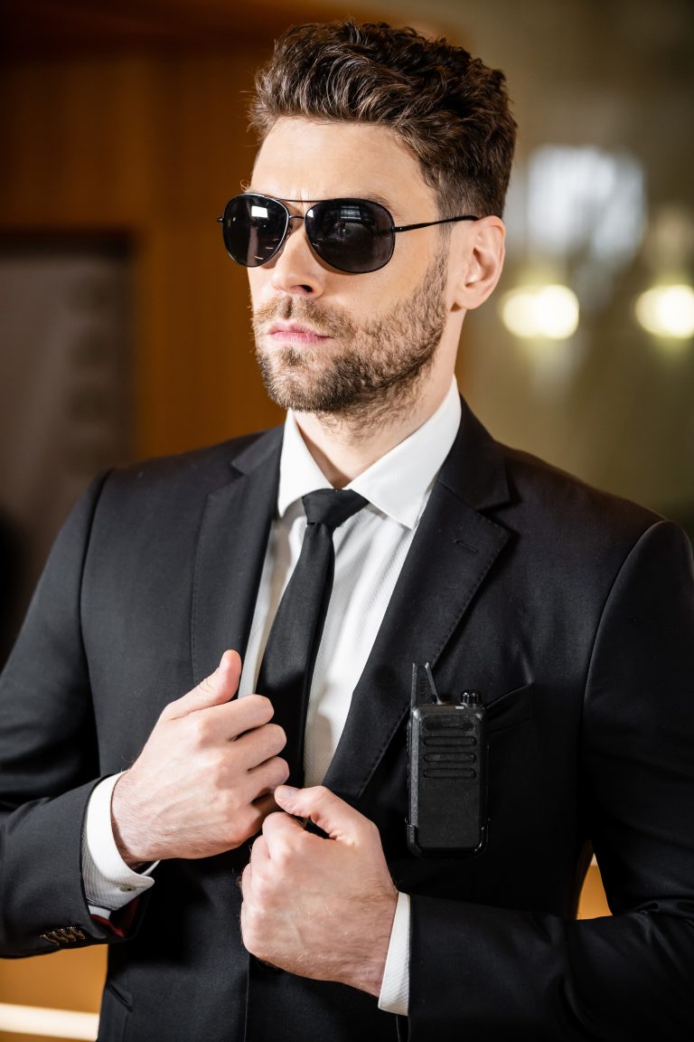 handsome bodyguard, security guard in suit with tie and sunglasses standing in hotel, professional headshots, radio transceiver attached to jacket pocket, bearded man working in hotel security