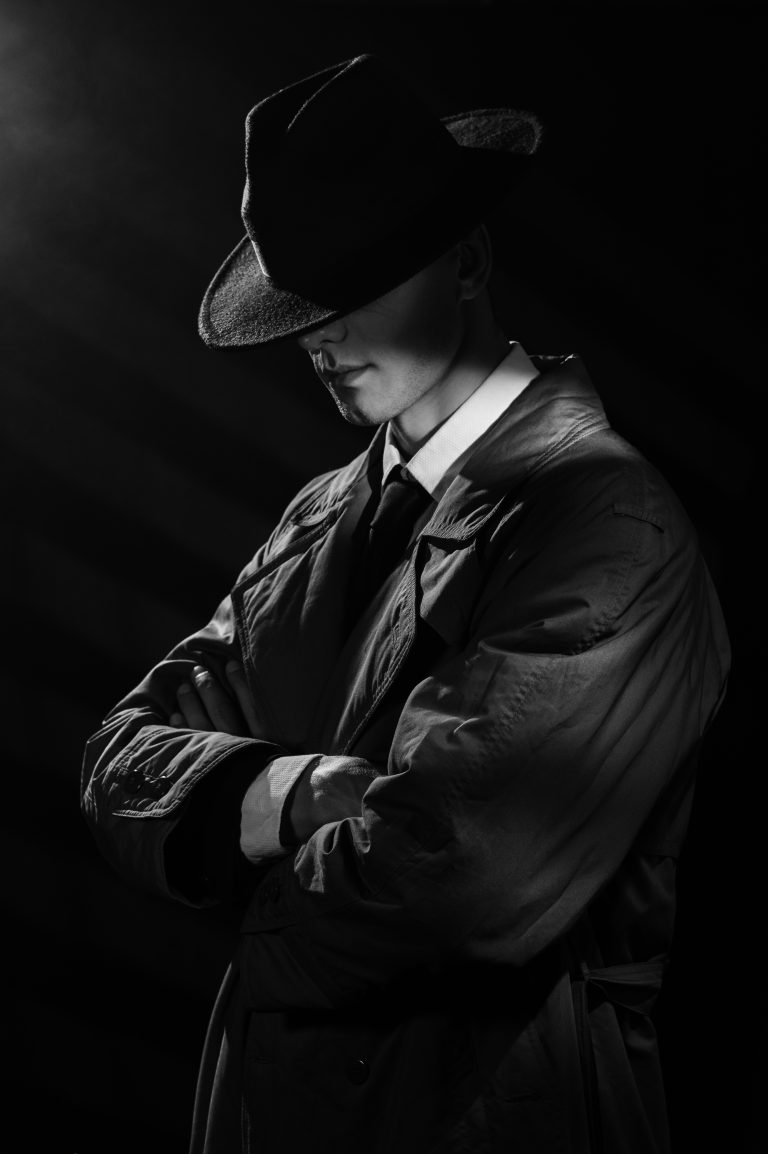 a dark silhouette of a man in a raincoat and hat with his head down in the style of crime noir. A dramatic noir portrait in the style of detectives of the 1950s.